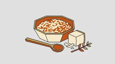 A bowl of rice and grains next to a wooden spoon, a block of tofu, and a small leafy branch.
